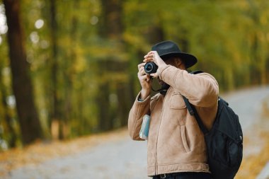 Genç siyahi bir adam ormanda elinde kamerayla dikiliyor. Erkek fotoğrafçı fotoğraf çekiyor. Kahverengi ceketli, siyah şapkalı ve sırt çantalı bir adam..