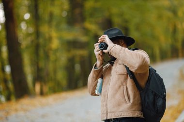 Genç siyahi bir adam ormanda elinde kamerayla dikiliyor. Erkek fotoğrafçı fotoğraf çekiyor. Kahverengi ceketli, siyah şapkalı ve sırt çantalı bir adam..
