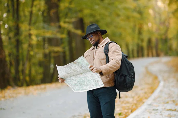 Young black man hitchhiking on road and looking at map. Male traveler feeling lost, traveling alone by autostop. Man wearing brown jacket, black hat and backpack.