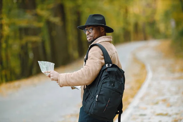 Young black man hitchhiking on road and looking at map. Male traveler feeling lost, traveling alone by autostop. Man wearing brown jacket, black hat and backpack.