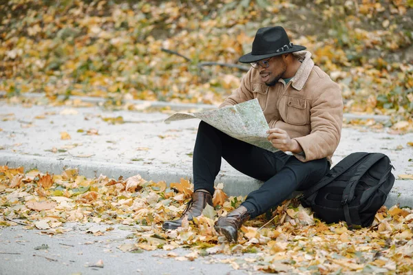 Young black man sitting on road and looking at map. Male traveler feeling lost, traveling alone by autostop. Man wearing brown jacket, black hat and backpack.