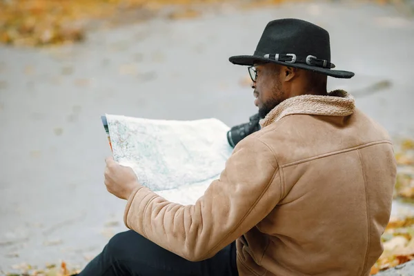 Young black man sitting on road in forest with a camera. Male photographer in a forest. Man wearing brown jacket, black hat and backpack.