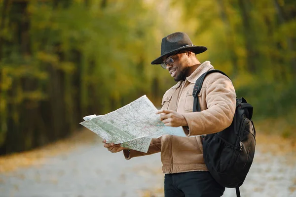 Young black man hitchhiking on road and looking at map. Male traveler feeling lost, traveling alone by autostop. Man wearing brown jacket, black hat and backpack.