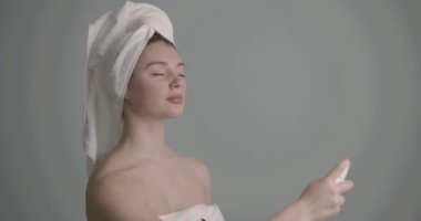 Kafasında havlu olan genç bir kadın yüz güzelliği ürünleri uyguluyor. Termal su ya da sprey içinde sis kullanmak. Scin care ürün reklamcılığı.