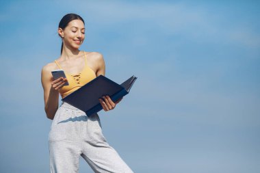Gökyüzü arka planında duran güzel kız. Sarı bluzlu kadın. Elinde belgeler olan bir kadın.
