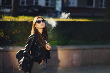 Şık bir kız şehirde yürürken e-sigara içiyor.