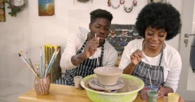 Couple smiling while doing creative painting on bowls in pottery workshop. Pottering Ideas, paint brushes.