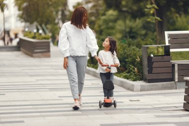 Anne ve kızı parkta scooter 'a biniyor. Çocuklar paten kaymayı öğreniyor. Küçük kız güneşli yaz gününde kayıyor.