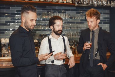 Meeting with the best friends. Three happy young men in casual wear talking and drinking beer while sitting in bar together. Man holding a phone in his hands.