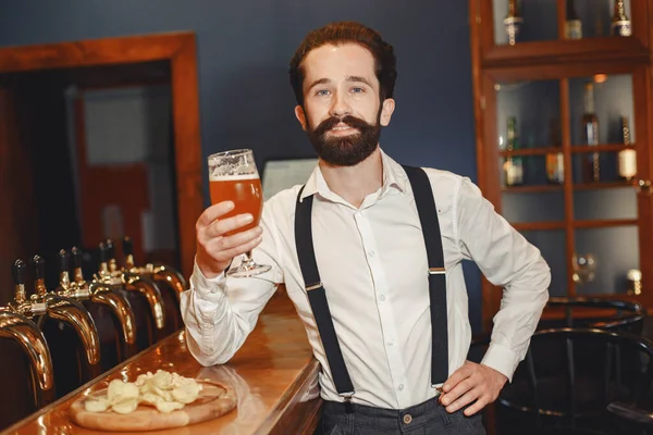 Attractive young man standing behind the bar. Bearded man drinking beer.