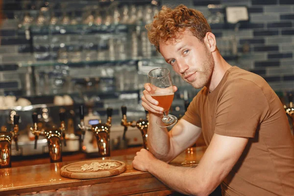 Attractive young man standing behind the bar. Man in a brown T-shirt holds a glass in his hands.