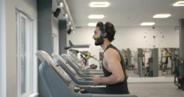 Side view of a young dark-haired athlete with a beard in a black sports uniform on a treadmill in a modern gym. Training concept