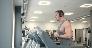 Side view of a young athlete in a gray T-shirt and headphones on a treadmill in a modern bright gym. Cardio training