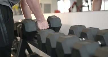 Close-up view of a young athlete in a gray T-shirt leaning over dumbbells in a modern bright gym. Selects the required free weight.