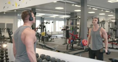 Side view of a young athlete in a gray T-shirt and headphones performing biceps exercises with dumbbells in front of a mirror in a modern bright gym. Training concept