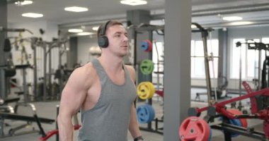 Side view of a young athlete in a gray T-shirt and headphones performing biceps exercises with dumbbells in front of a mirror in a modern bright gym. Training concept
