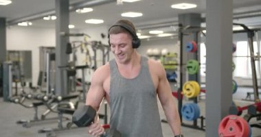 Front view of a young athlete in a gray T-shirt and headphones performing biceps exercises with dumbbells in front of a mirror in a modern bright gym. Training concept