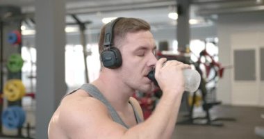 Side view of a young athlete in a gray T-shirt and headphones drinking water and wiping with a towel in a modern bright gym. Break between exercises. Training concept