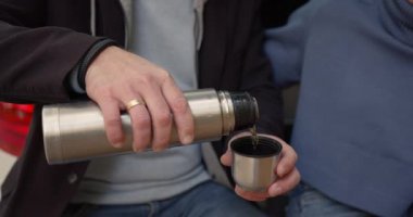 Close-up view of a young man pouring a cup of coffee from a thermos. Autumn date in nature
