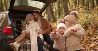 In the background, young parents are sitting in the trunk of a car. In front of the kids in hats with pompoms smiling. Autumn family leisure