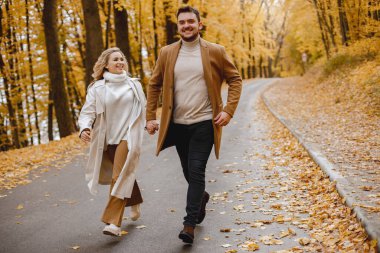 Young man and woman walking outside wearing beige coats. Blond woman and brunette man in autumn forest. Romantic couple holding hands.