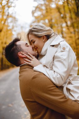 Young man and woman walking outside wearing beige coats. Blond woman and brunette man in autumn forest. Romantic couple kissing.