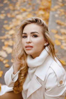 Young woman sitting on a curb in autumn forest. Blonde woman touching her hair. Girl wearing beige coat and brown trousers.