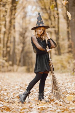 Blonde young girl witch in forest on Halloween. Girl wearing black dress and cone hat. Witch holding a broom.