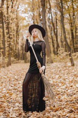 Blonde young girl witch in forest on Halloween. Girl wearing black dress and cone hat. Witch holding a broom.
