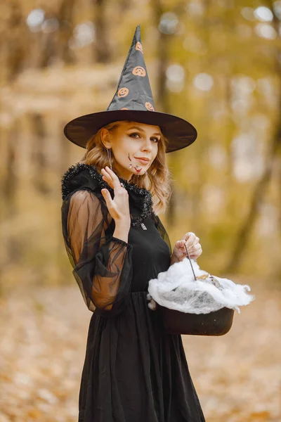 Blonde young girl witch in forest on Halloween. Girl wearing black dress and cone hat. Witch holding a magician stuff.