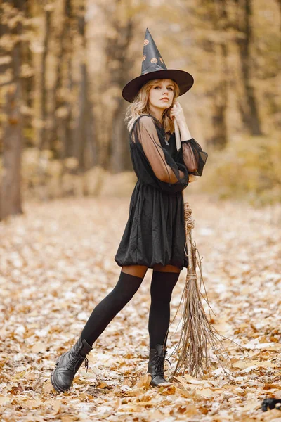 Blonde young girl witch in forest on Halloween. Girl wearing black dress and cone hat. Witch holding a broom.