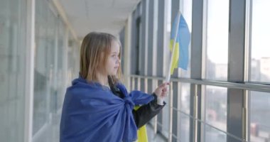 Little Ukrainian girl with a national yellow-blue flag. She is standing inside the glass bulding and showing Ukrainianf flag. Russian agression concept.