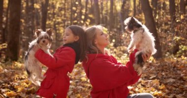 Young mother and daughter sit down and hold dogs in their arms. Warm up with animals on the lawn on a sunny autumn day.