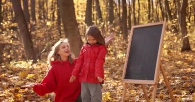 Mother and daughter in red sat down at a board in the autumn forest. They play with leaves and smile. Happy leisure