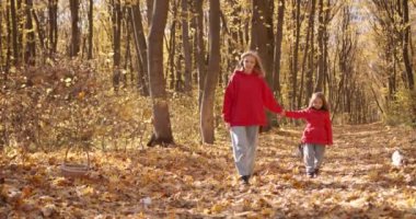 A mother in a red jacket holds her daughters hand. They smile and walk through the autumn park. Happy family leisure