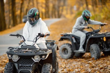 Focus on a woman riding an ATV quad bikes in autumn forest. Two friends maneuvering off-road ATV. Couple wearing grey sportive costumes.