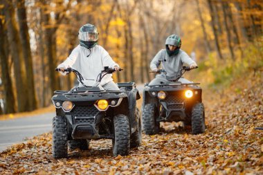 A young caucasian man and woman riding an ATV quad bikes in autumn forest. Two friends maneuvering off-road ATV. Couple wearing grey sportive costumes.