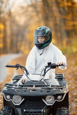A young caucasian woman in helmet riding an ATV quad bike in autumn forest. Girl maneuvering off-road ATV. Woman wearing grey sportive costume.