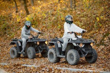 A young caucasian man and woman riding an ATV quad bikes in autumn forest. Two friends maneuvering off-road ATV. Couple wearing grey sportive costumes.