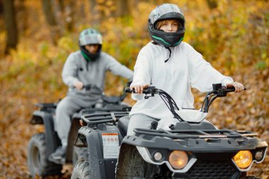 A young caucasian man and woman riding an ATV quad bikes in autumn forest. Two friends maneuvering off-road ATV. Couple wearing grey sportive costumes.