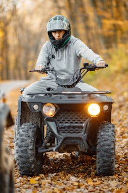 A young caucasian man in helmet riding an ATV quad bike in autumn forest. Boy maneuvering off-road ATV. Man wearing grey sportive costume.