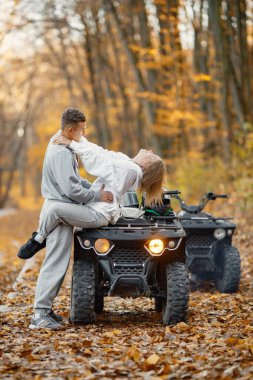 A young caucasian man and woman riding an ATV quad bike in autumn forest. Lovely couple hugging. Couple wearing grey sportive costumes.