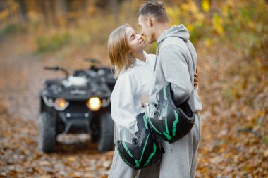 Young caucasian man and woman near an ATV quad bike in autumn forest. Lovely couple kissing while holding a helmets in hands. Couple wearing grey sportive costumes.