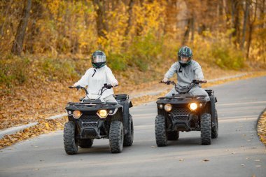 A young caucasian man and woman riding an ATV quad bikes in autumn forest. Couple maneuvering off-road ATV. Couple wearing grey sportive costumes.