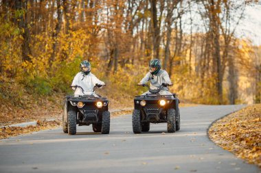 A young caucasian man and woman riding an ATV quad bikes in autumn forest. Couple maneuvering off-road ATV. Couple wearing grey sportive costumes.