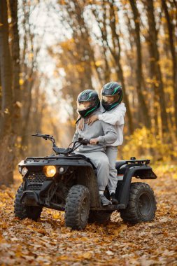 A young caucasian man and woman riding an ATV quad bike in autumn forest. Couple maneuvering off-road ATV. Couple wearing grey sportive costumes.