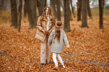 Young woman and little girl in autumn forest. Woman and her daughter throwing dried leaves in the air. Girl wearing fashion grey dress with a jacket.
