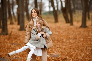Young woman and little girl in autumn forest. Woman holding her daughter on hands and hugging. Girl wearing fashion grey dress with a jacket.