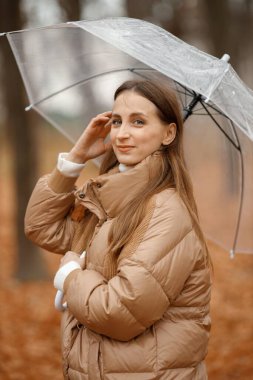 Young woman standing in autumn forest. Brunette woman holding a transparent umbrella. Girl wearing fashion brown jacket.