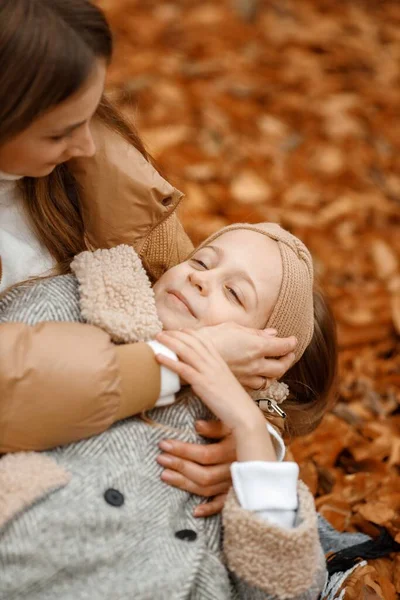 Little girl in fashion clothes laying on mothers lap in autumn forest. Girl close her eyes. Girl wearing grey coat and a headband.